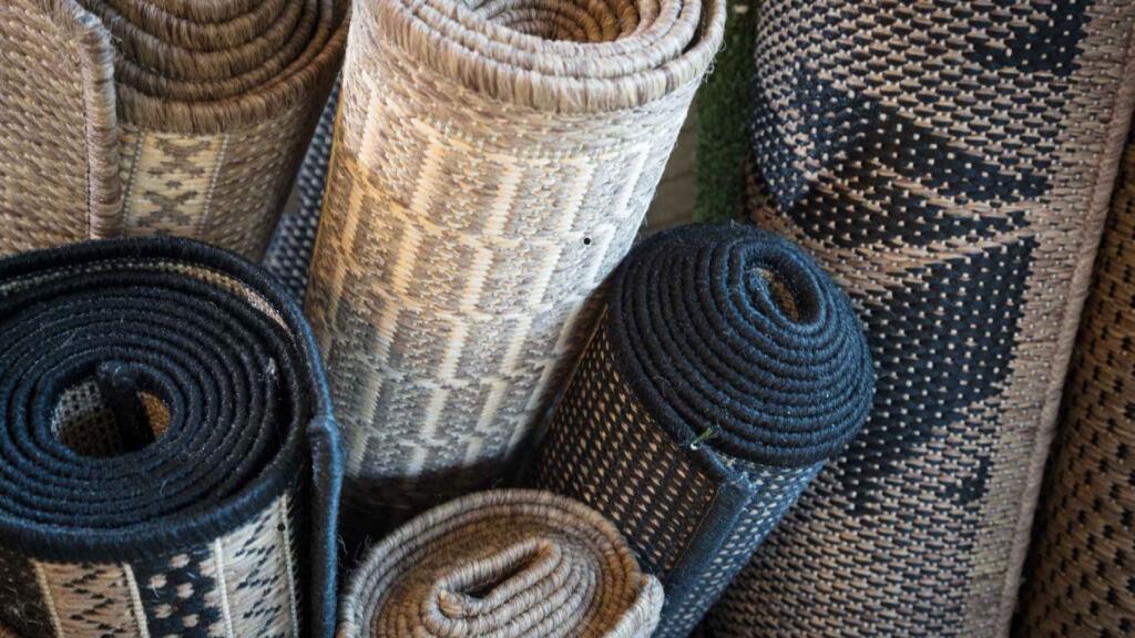 The image shows several rolls of woven rugs stacked closely together. The rugs come in various shades of neutral colors like beige, black, and brown, and feature different patterns, including geometric designs and woven textures. The materials appear to be sturdy and rough, likely made from natural fibers such as jute or sisal. The rolls are tightly wound, showcasing the ends of the rugs prominently. The overall setting gives a sense of craftsmanship and durability, with the patterns adding a touch of aesthetic appeal.
