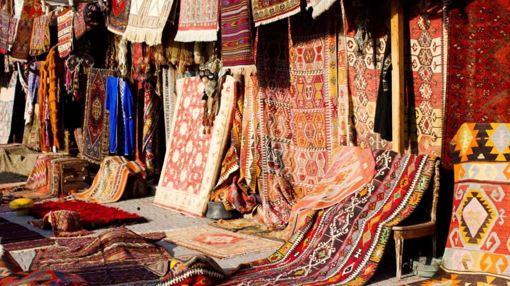 A vibrant outdoor market stall displaying an array of colorful rugs and textiles. The rugs feature intricate geometric and floral patterns in rich tones of red, orange, and brown, with a variety of textures. Some rugs are hanging, while others are laid out or draped over chairs. The scene is sunlit, showcasing the craftsmanship and cultural heritage of the handmade rugs, likely in a traditional marketplace setting.
