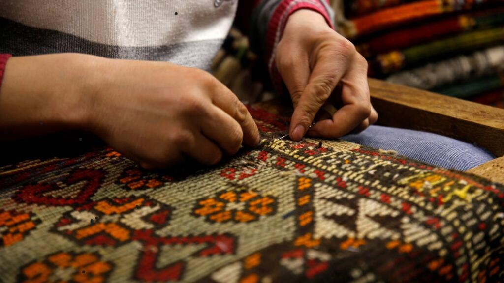 A close-up of a person's hands meticulously working on a colorful woven rug. The hands are using a needle or small tool to stitch or repair the intricate geometric and floral patterns in shades of red, orange, and white. The scene emphasizes the detailed craftsmanship and skill involved in traditional rug-making, highlighting the artisan's careful attention to the design and texture of the fabric.
