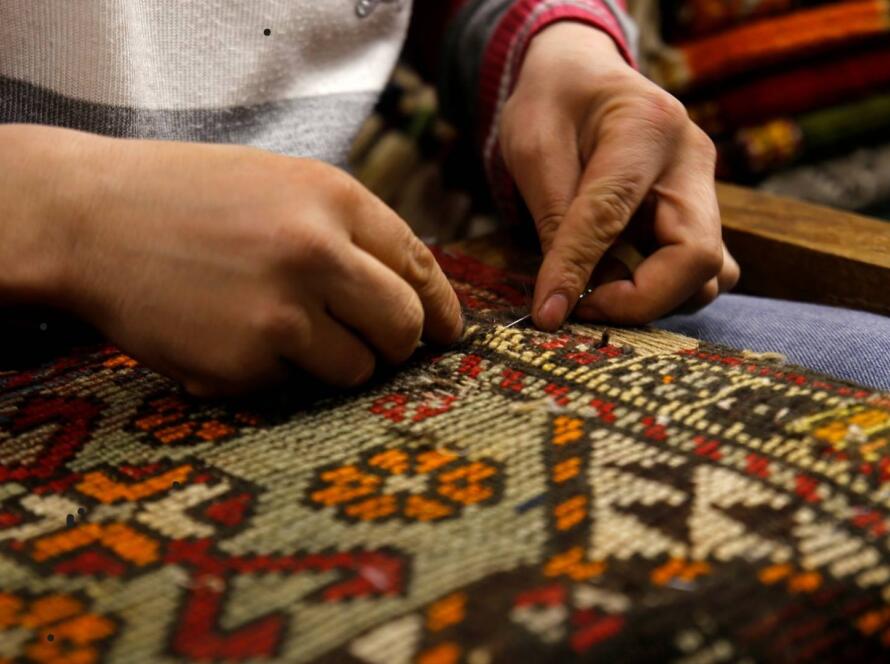 A close-up of a person's hands meticulously working on a colorful woven rug. The hands are using a needle or small tool to stitch or repair the intricate geometric and floral patterns in shades of red, orange, and white. The scene emphasizes the detailed craftsmanship and skill involved in traditional rug-making, highlighting the artisan's careful attention to the design and texture of the fabric.