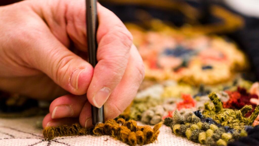 A close-up of a person's hand using a tool to work on a detailed rug or textile piece. The hand is pushing yarn or fiber through the fabric, creating a textured design with loops of yarn in various colors, such as brown, green, and red. The image highlights the precision and skill involved in the rug-making process, focusing on the intricacies of creating patterns and textures by hand.
