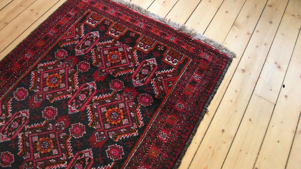 A traditional red and black rug with intricate geometric patterns, placed on a polished light wooden floor. The rug features bold designs with floral and diamond-shaped motifs, surrounded by a detailed border. The vibrant colors and craftsmanship of the rug stand out against the light wood, creating a warm and elegant home interior setting.







