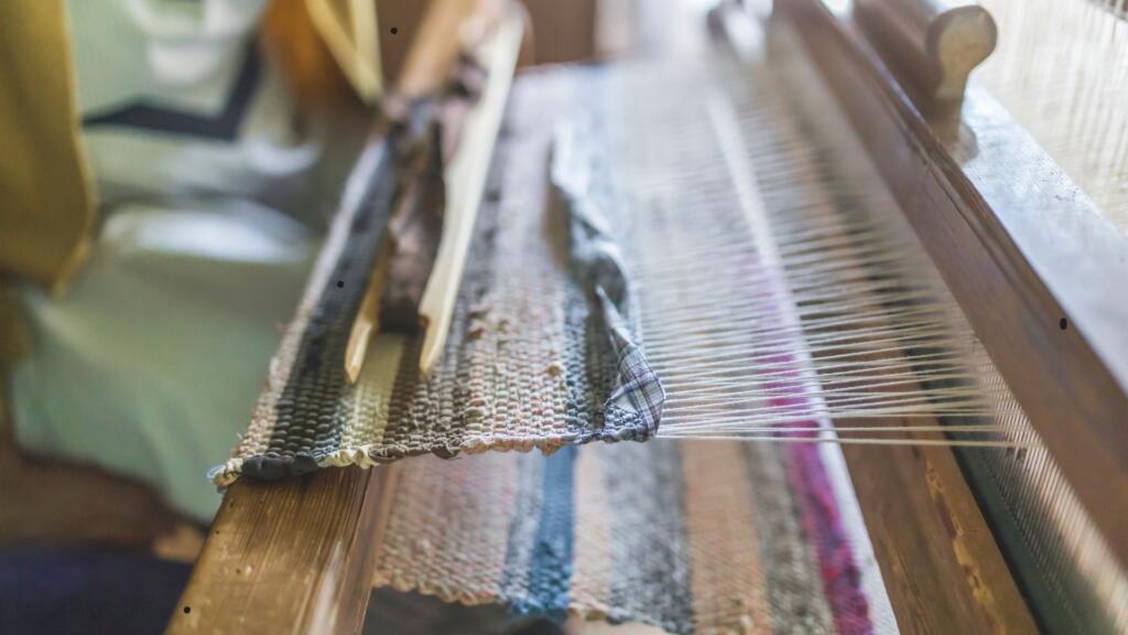 The image shows a close-up view of a traditional handloom with a partially woven textile. The wooden loom has threads stretched across it, with a shuttle carrying the weft thread visible on top. The fabric being woven displays a mix of colors and textures, giving a rustic, handcrafted feel. The background is softly blurred, emphasizing the focus on the loom and textile in progress.






