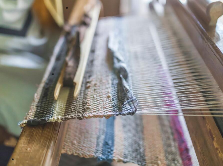 The image shows a close-up view of a traditional handloom with a partially woven textile. The wooden loom has threads stretched across it, with a shuttle carrying the weft thread visible on top. The fabric being woven displays a mix of colors and textures, giving a rustic, handcrafted feel. The background is softly blurred, emphasizing the focus on the loom and textile in progress.