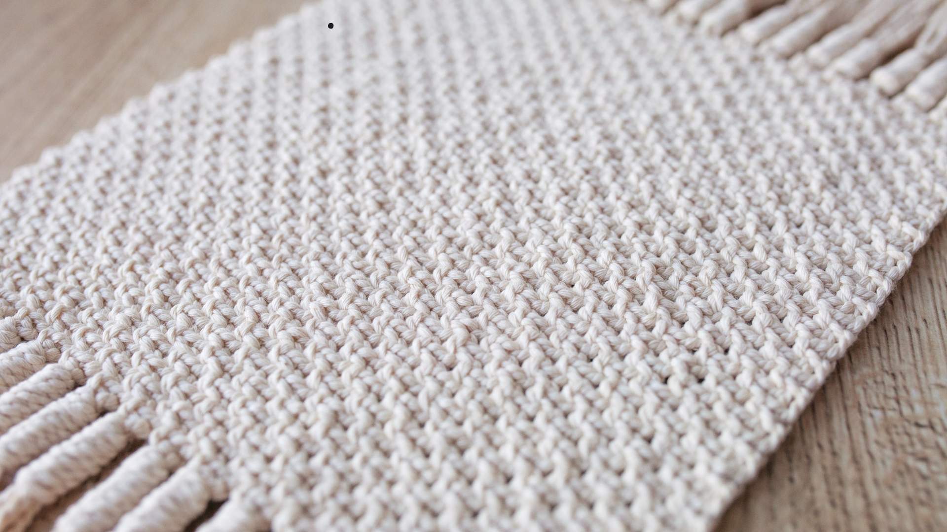 The image shows a close-up of a woven textile in a natural cream color, featuring a uniform, textured weave with fringes on the edge. It rests on a wooden surface, emphasizing its handmade, minimalist style.