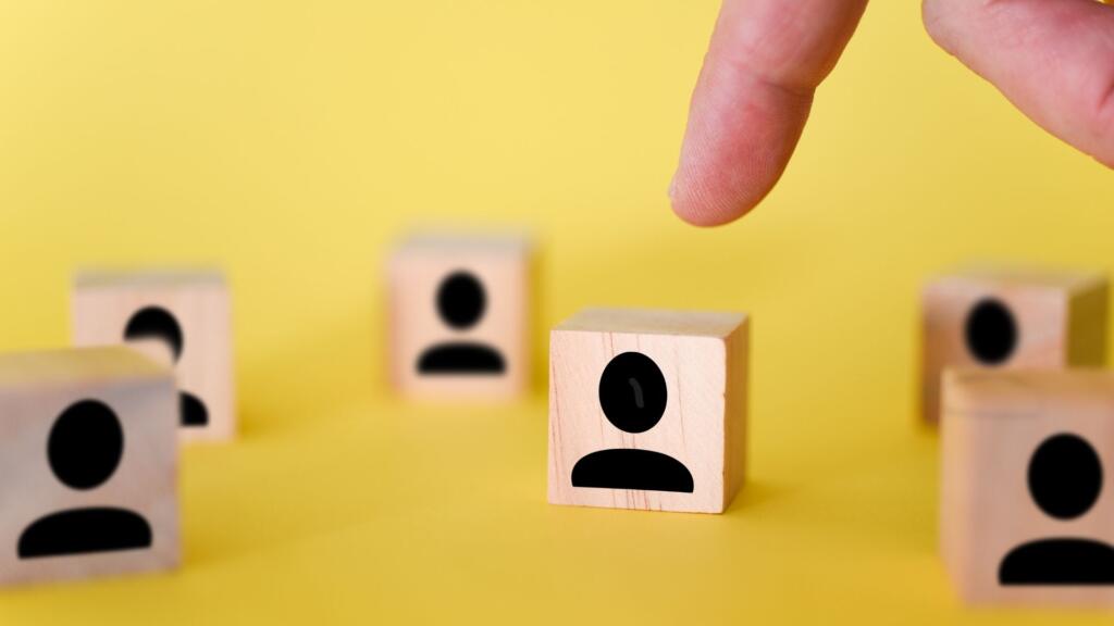 The image shows a hand reaching toward a wooden block with a simple icon of a person on it. The block is one of several placed on a bright yellow background. Each wooden block has the same person icon, resembling a stylized human silhouette, in black. The scene conveys the concept of selection, recruitment, or decision-making, with the hand appearing as if it's about to pick one of the blocks, suggesting a focus on individuals or choices within a group. The overall aesthetic is minimalistic and clean.
