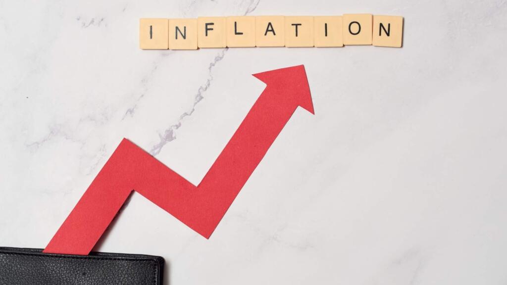 The image shows the word "INFLATION" spelled out with Scrabble-like letter tiles on a light marble surface. Below the word, there is a bold red arrow pointing upward at an angle, indicating a rise, symbolizing the concept of increasing inflation. The base of the arrow seems to emerge from a black wallet at the bottom left corner, visually linking inflation to financial impact or cost of living. The clean design and contrasting elements emphasize the upward movement associated with inflation.
