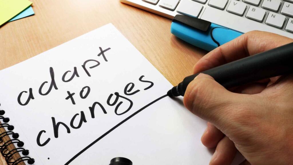 The image shows a hand holding a black marker writing the words "adapt to changes" on a spiral-bound notebook. The text is handwritten in bold, clear letters. The scene takes place on a wooden desk, with a keyboard and some stationery visible in the background, including sticky notes and a blue highlighter. The image conveys a message of flexibility, growth, or adjustment, emphasizing the importance of adapting to new circumstances or changes. The casual, handwritten style adds a personal and motivational touch to the message.
