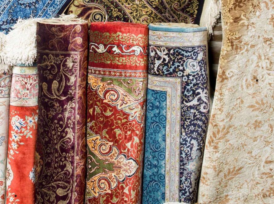 The image shows a variety of intricately patterned, colorful rugs rolled up and displayed. The rugs feature detailed floral and ornamental designs in rich colors such as red, blue, gold, and purple. Some of the rugs are partially unrolled, revealing fringed edges, while others remain tightly wound. The textures and patterns suggest a focus on traditional craftsmanship, with a mix of vibrant and muted tones. The rugs are likely made from high-quality materials, giving the display a luxurious and artistic feel.