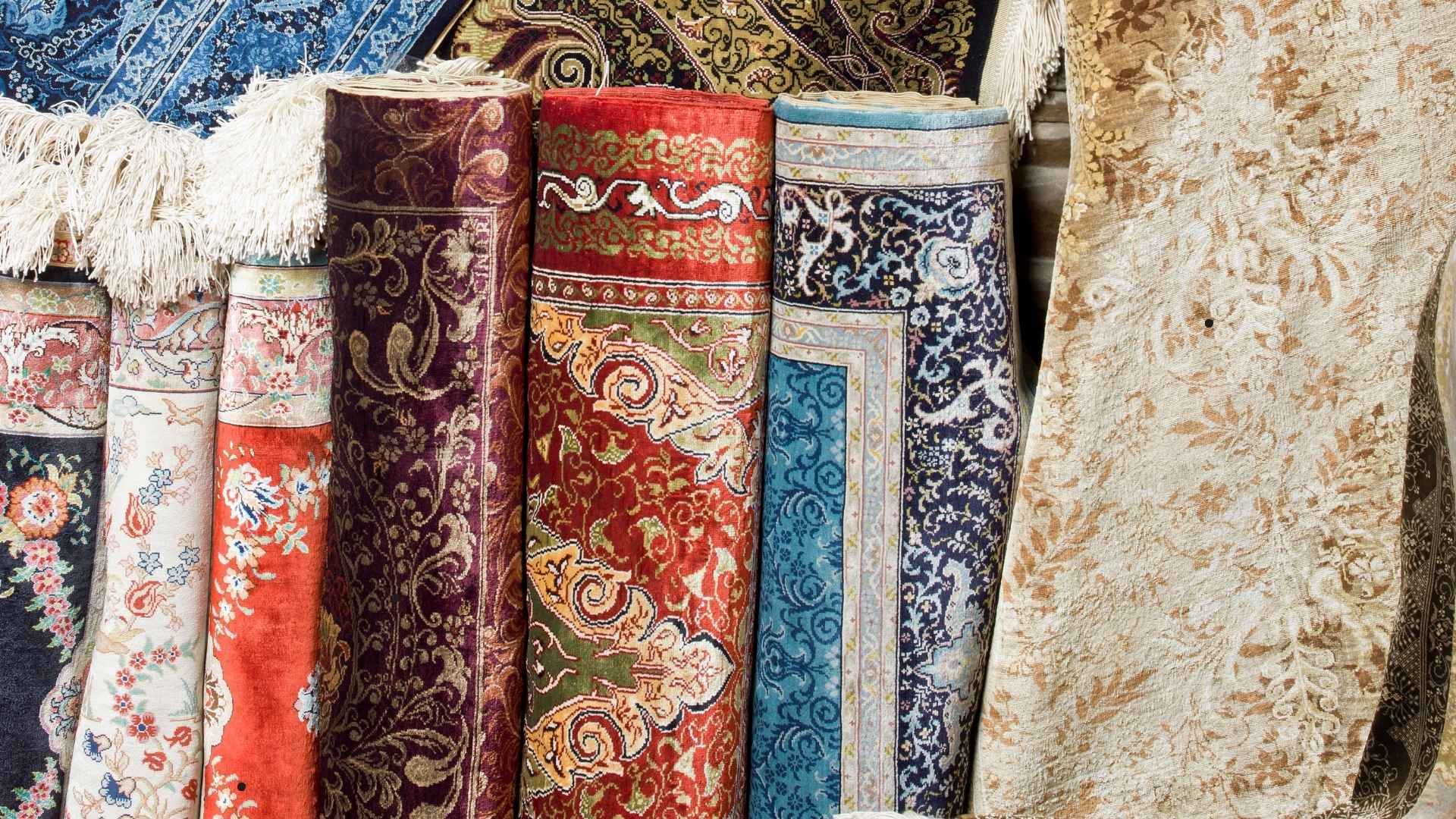 The image shows a variety of intricately patterned, colorful rugs rolled up and displayed. The rugs feature detailed floral and ornamental designs in rich colors such as red, blue, gold, and purple. Some of the rugs are partially unrolled, revealing fringed edges, while others remain tightly wound. The textures and patterns suggest a focus on traditional craftsmanship, with a mix of vibrant and muted tones. The rugs are likely made from high-quality materials, giving the display a luxurious and artistic feel.






