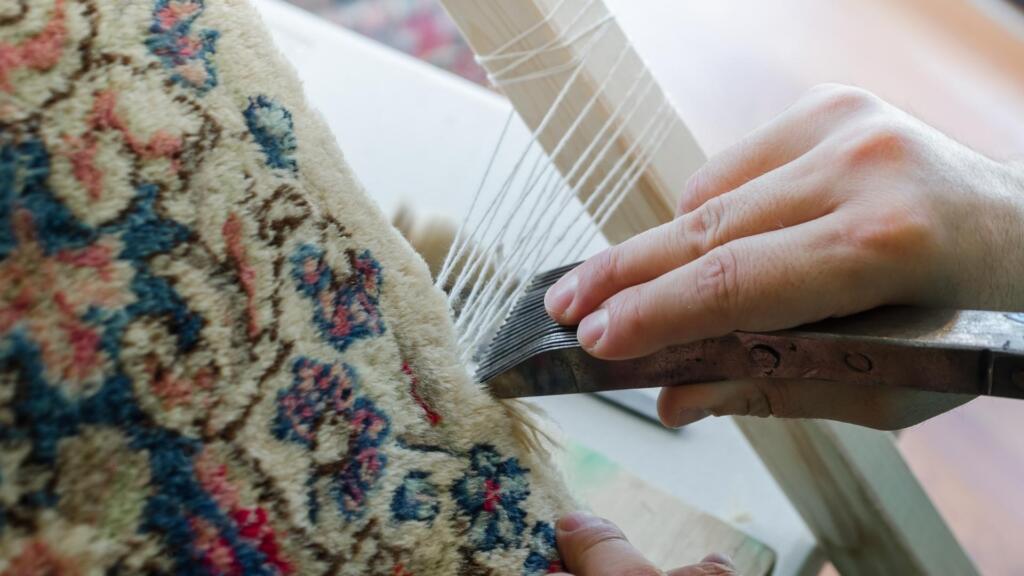 The image shows a close-up of hands working on a traditional rug weaving process. One hand holds a metal tool, possibly a comb or beater, used to tighten and align the fibers on a loom. The other hand stabilizes the fabric, which displays a floral or ornate pattern in colors like blue, red, and beige. White threads from the loom are visible, suggesting the rug is being handwoven. The image highlights the craftsmanship and detail involved in rug-making, emphasizing the use of traditional techniques and materials.
