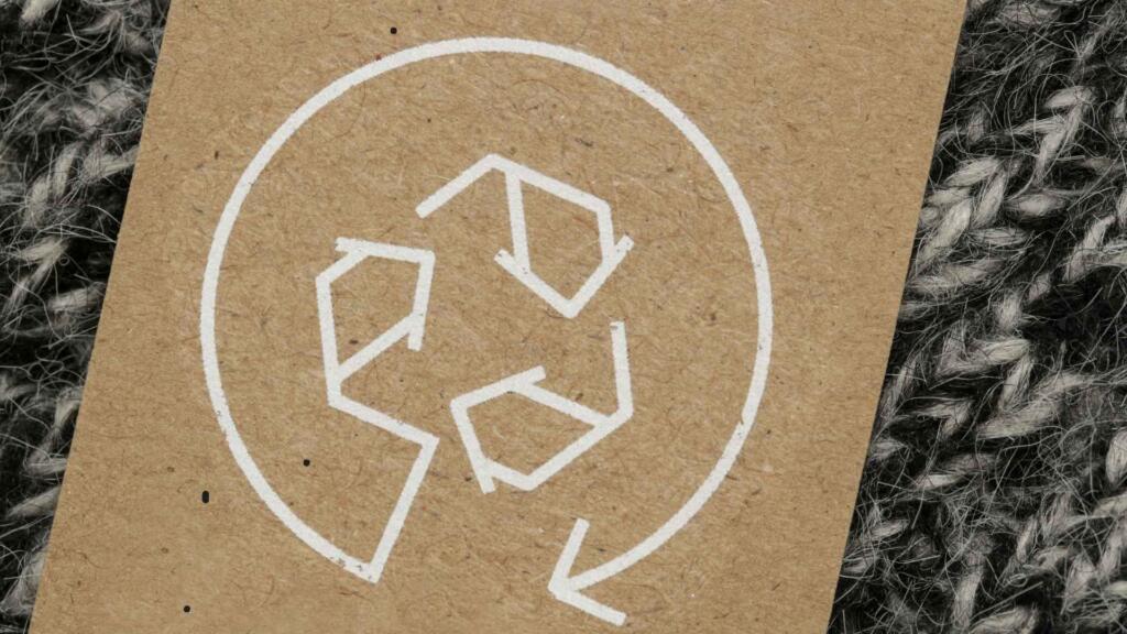 A brown cardboard tag with a white recycling symbol printed on it, placed on a textured gray and black woven fabric. The image emphasizes eco-friendly and sustainable practices, with the recycling logo symbolizing environmental responsibility and material reuse in the textile industry.
