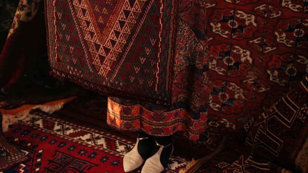 A person standing among a display of intricately patterned rugs, with their feet visible in white shoes. The rugs are richly colored, featuring deep reds, browns, and geometric designs. The scene highlights the traditional craftsmanship and artistic detail of the rugs, with a warm and inviting ambiance created by the rich tones and textures of the woven fabric.
