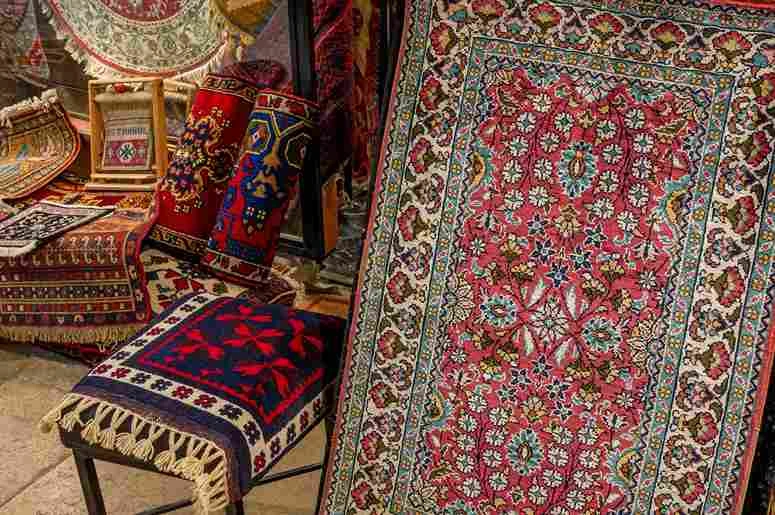 A vibrant display of traditional rugs in a market setting, featuring a prominent rug with intricate floral and geometric patterns in red, blue, and beige tones. Smaller, colorful rugs with detailed designs are arranged nearby, showcasing the rich craftsmanship and cultural artistry typical of handcrafted textiles.