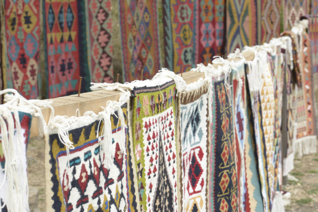 A row of colorful handwoven rugs with intricate geometric patterns hanging outdoors, showcasing traditional craftsmanship and vibrant cultural designs.