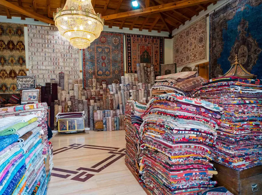 An interior view of a rug store showcasing stacks of vibrant, folded rugs and rolled-up carpets in a variety of colors and patterns. The walls are adorned with large, intricately designed rugs, and a chandelier hangs from the wooden ceiling, adding to the traditional and artistic ambiance of the space.