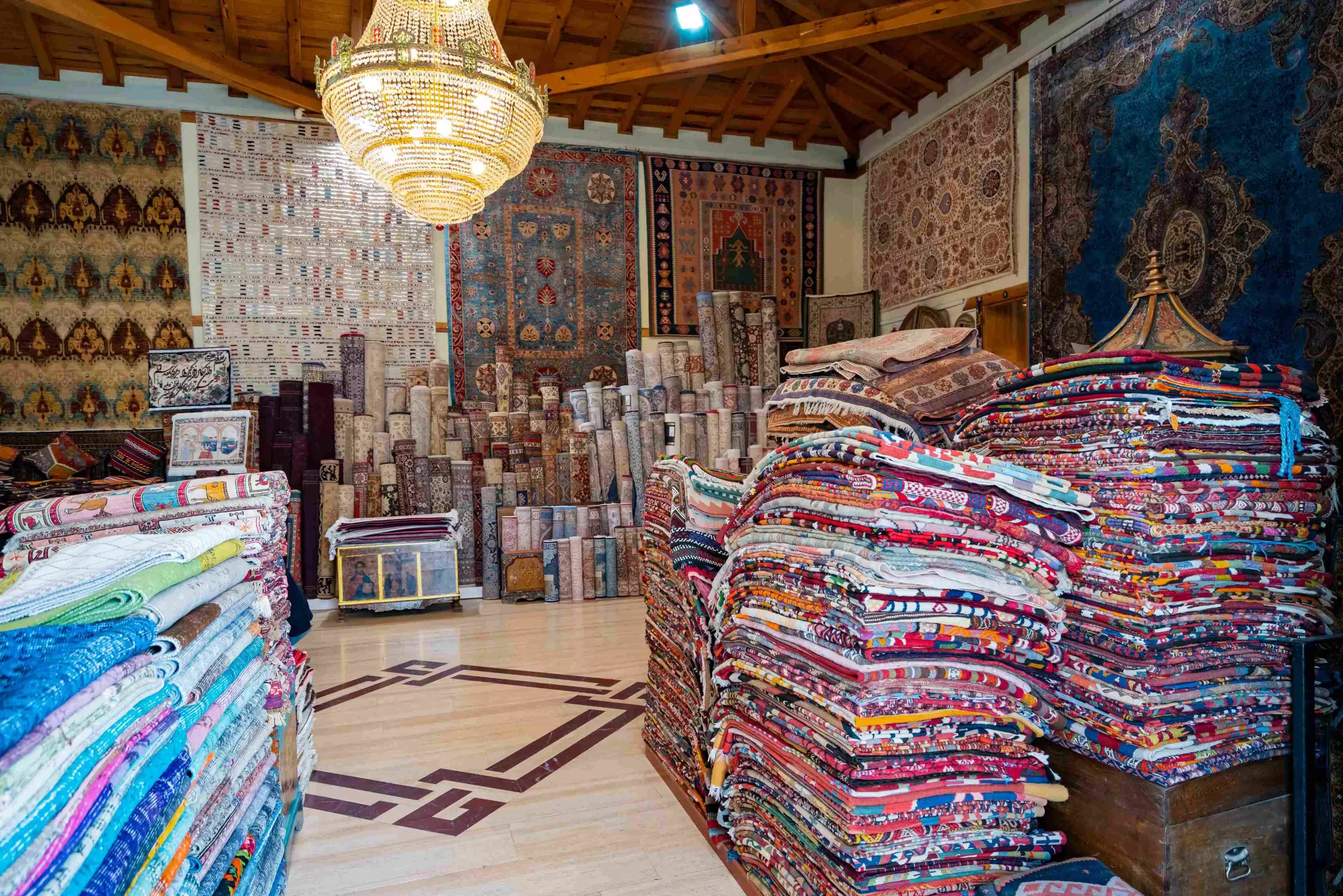 An interior view of a rug store showcasing stacks of vibrant, folded rugs and rolled-up carpets in a variety of colors and patterns. The walls are adorned with large, intricately designed rugs, and a chandelier hangs from the wooden ceiling, adding to the traditional and artistic ambiance of the space.