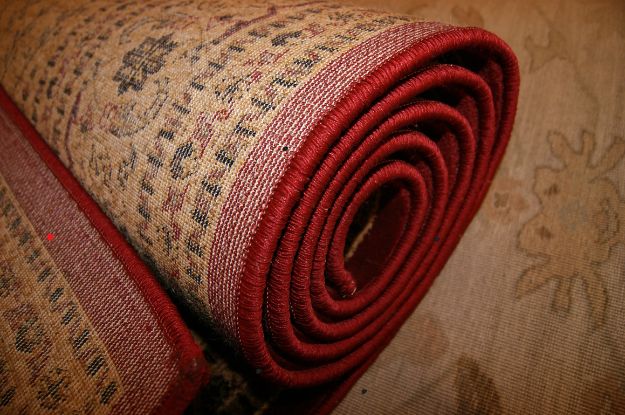 A close-up image of a rolled traditional rug with intricate patterns and a red border. The rug features detailed floral designs and rich textures, showcasing high-quality craftsmanship and durable materials.