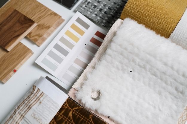 A selection of interior design materials, including fabric swatches, wood samples, and a color palette card with neutral tones. The items are arranged neatly, showcasing a variety of textures, such as soft fur-like fabric and woven patterns, along with light wood samples. This display represents the process of coordinating materials for home decor or renovation projects.
