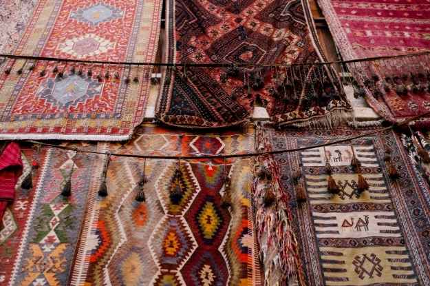 Display of traditional, colorful rugs featuring intricate geometric and tribal patterns, hanging side by side in a market setting