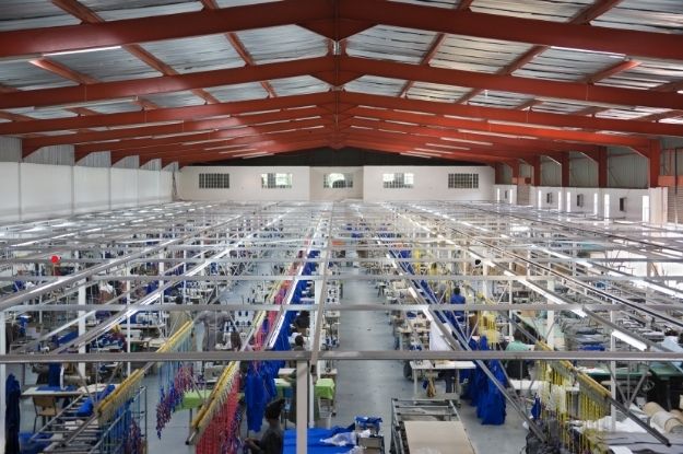 Spacious rug manufacturing facility with rows of equipment and materials, showing the industrial production process under a high, red-beamed roof.