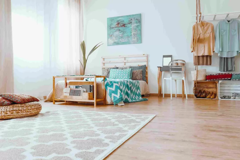 A bright and cozy bedroom with minimalist decor, featuring a light wood bed adorned with patterned pillows and a teal blanket. A soft beige and white rug with a geometric pattern covers the wooden floor, adding warmth to the space. The room includes a woven floor cushion, neatly hung clothing, and a small work area, creating a relaxing and organized atmosphere.
