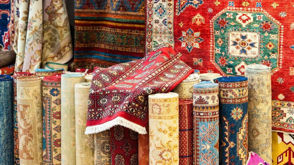 A vibrant collection of rolled-up and hanging traditional rugs with intricate patterns in various colors, showcasing a wide range of handcrafted designs.