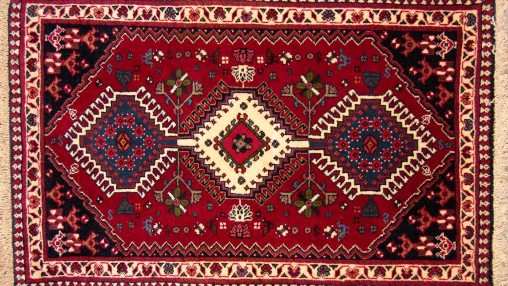 A traditional Persian-style rug with intricate geometric patterns and vibrant red, blue, and cream colors, featuring floral motifs and detailed border designs.