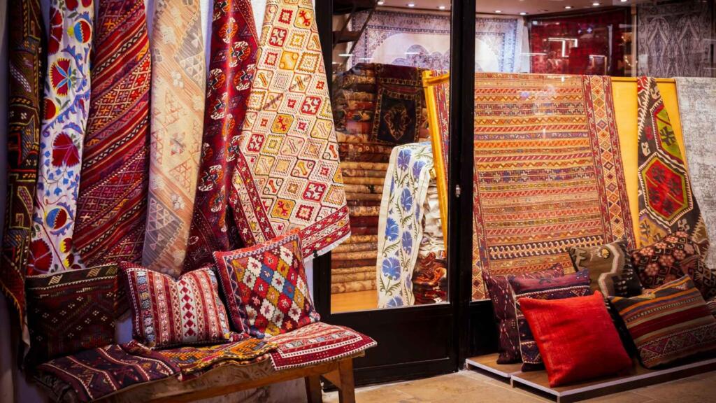 A variety of colorful traditional rugs and cushions displayed in a shop, showcasing intricate patterns and craftsmanship, creating a cozy and vibrant atmosphere.