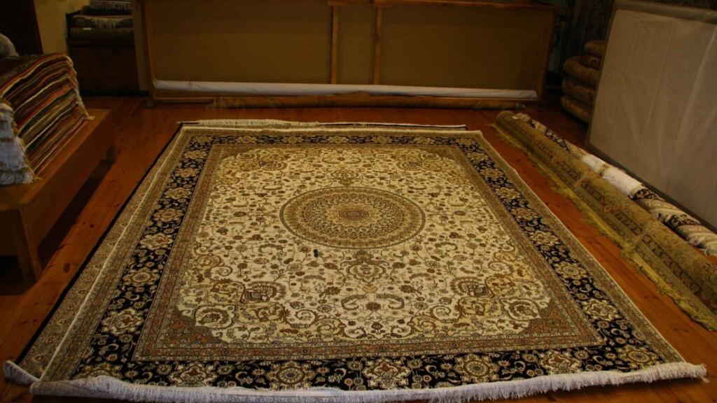 An ornate, traditional Persian rug with intricate floral motifs and an elegant medallion design, displayed on a wooden showroom floor, showcasing fine craftsmanship.