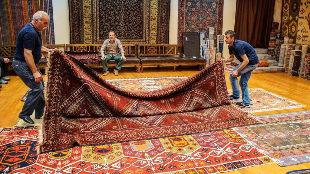 Two men unfolding a traditional red patterned rug in a rug showroom, showcasing the variety of colorful handmade rugs available for display and purchase.