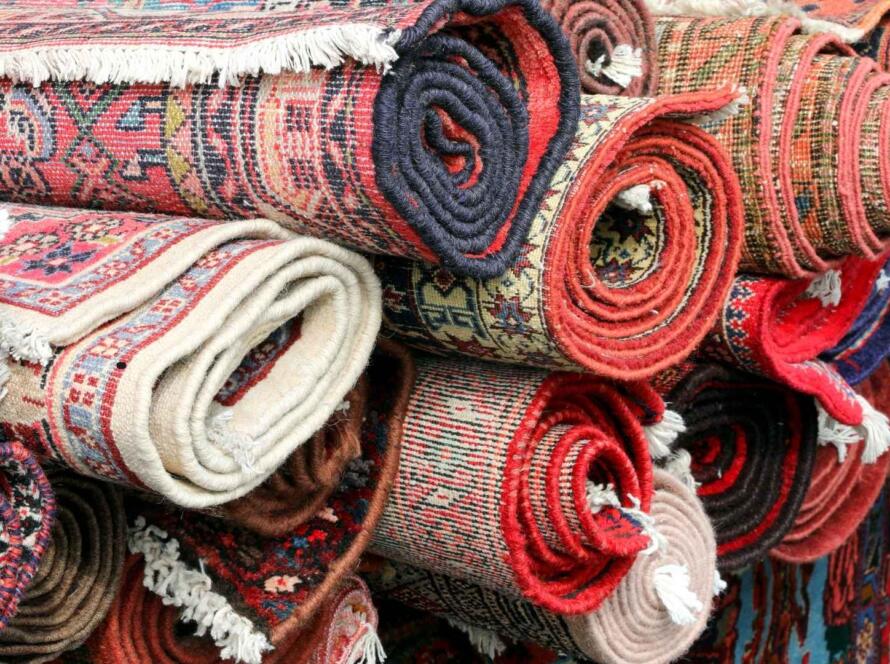 A stack of rolled-up traditional rugs in various vibrant colors and intricate patterns, showcasing the craftsmanship and diversity of handmade textiles.