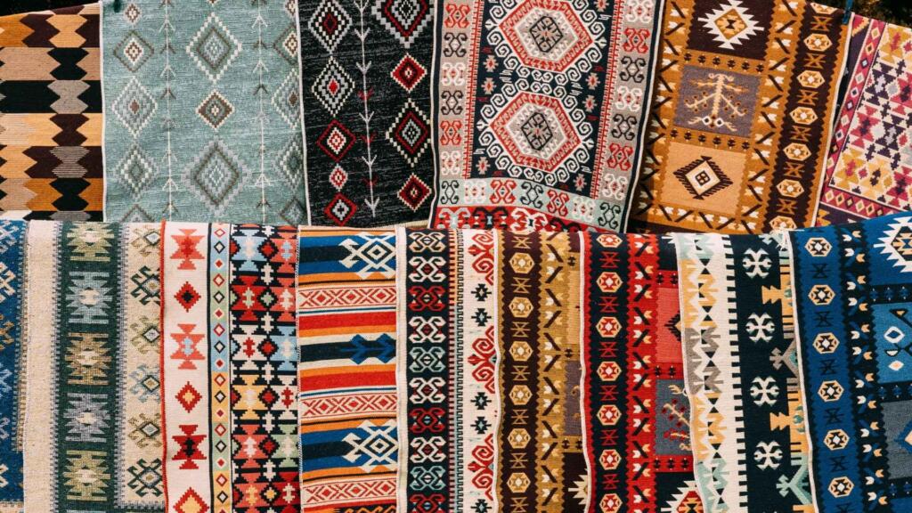 A colorful assortment of traditional rugs with intricate geometric patterns, showcasing a variety of vibrant designs and textures in a market display.
