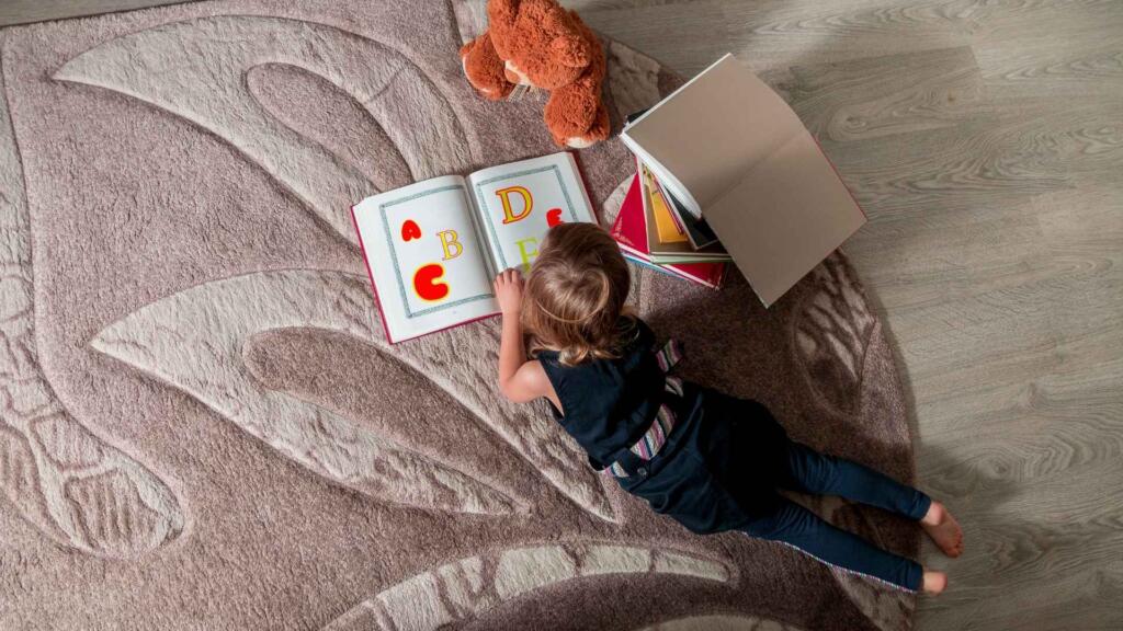 A young child lying on a cozy, patterned rug while reading an alphabet book, with a teddy bear and a stack of books nearby, showcasing a comfortable and playful learning environment.