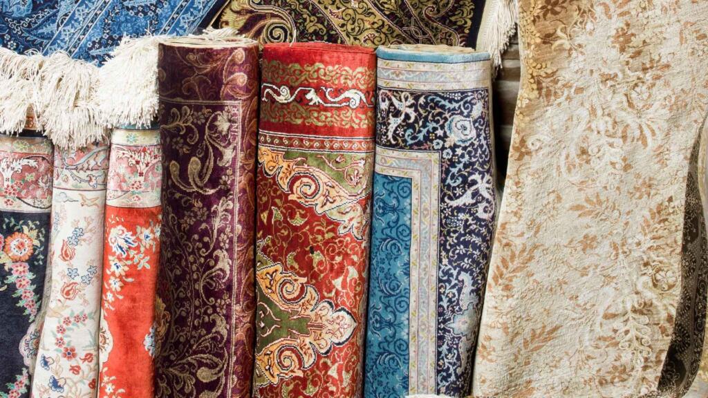 A collection of rolled-up, intricately patterned rugs in vibrant colors, showcasing various traditional designs and luxurious textures.