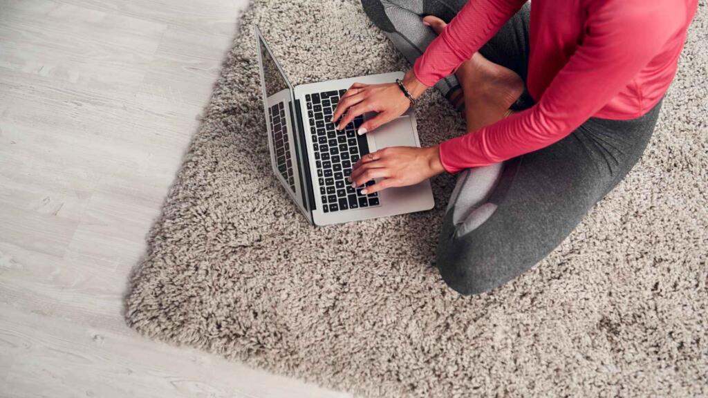 Woman shopping for rugs online while sitting comfortably on a plush, textured rug, emphasizing the convenience of e-commerce for home decor products.