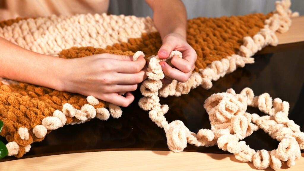 A person knitting a chunky knit blanket with yarn in a brown and cream color.