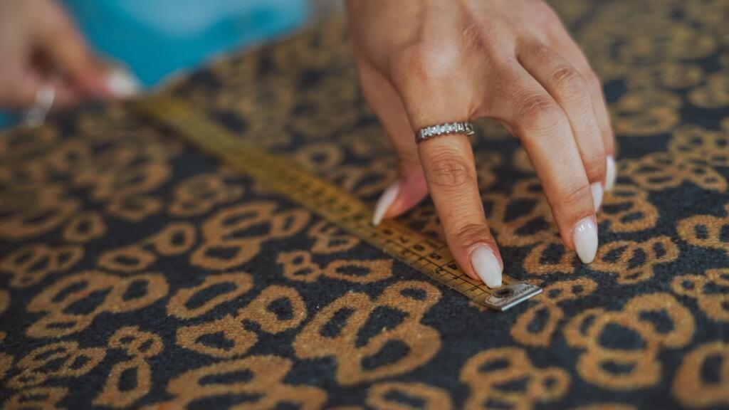 An image showing the process of measuring a room or space for a custom rug. A person is using a tape measure to carefully measure the length and width of the area where the rug will be placed. The floor is visible, and the measurement marks are being recorded with a notepad or device.