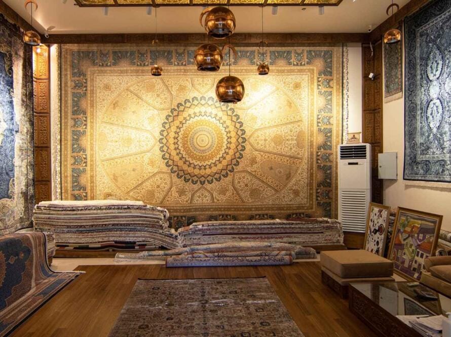 "A luxurious display of handmade area rugs featuring intricate patterns in a showroom setting, with rugs adorning the walls and stacked neatly on the floor."