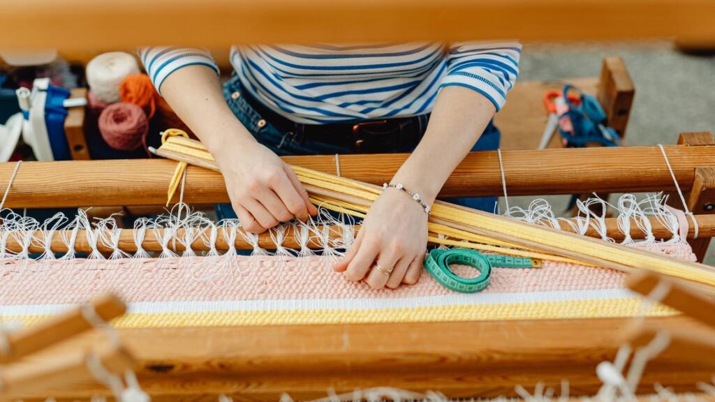 "Artisan weaving an Indian carpet on a traditional wooden loom with threads and measuring tools in view."