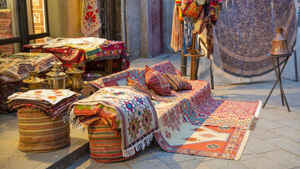 An outdoor display of colorful custom shape carpets and traditional textiles arranged on stools and benches, creating a vibrant and inviting scene.