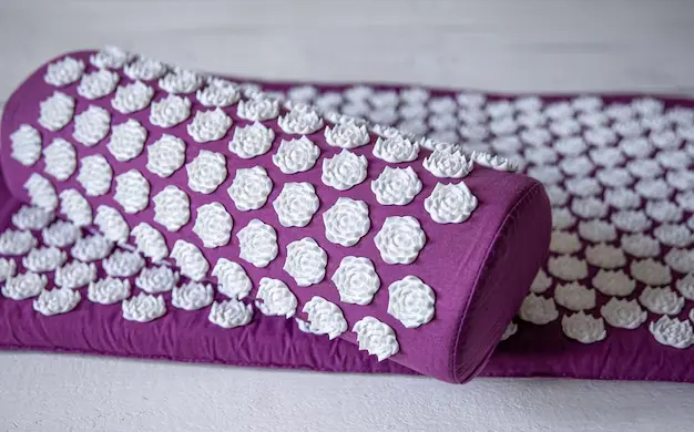 A purple acupressure mat and pillow set covered with white, lotus-shaped massage points, designed for relaxation and pain relief, placed on a white surface.