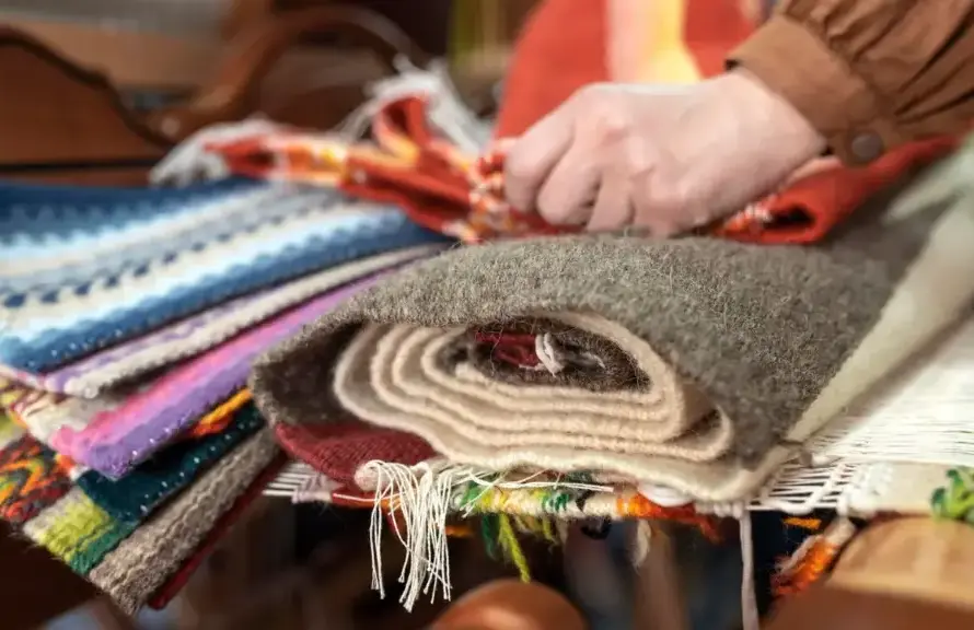 A person arranging colorful, stacked fabric rugs, including woolen and woven textures, on a wooden table.
