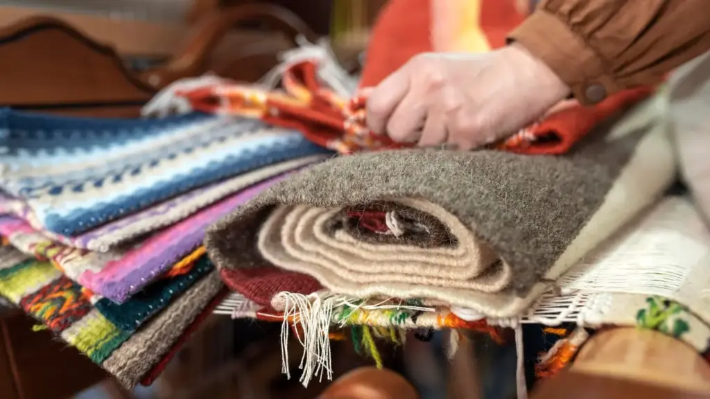 A person arranging colorful, stacked fabric rugs, including woolen and woven textures, on a wooden table.