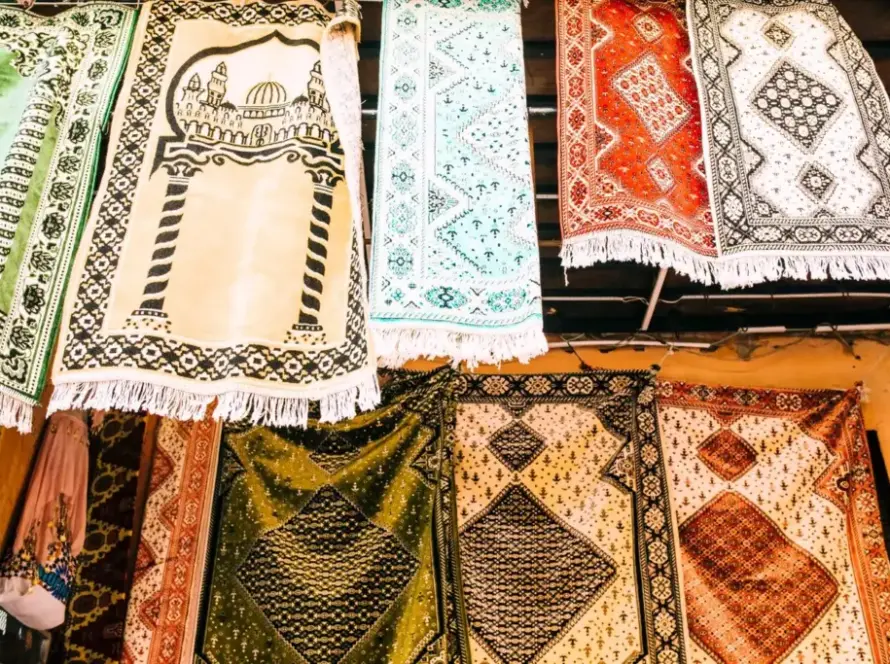 A collection of colorful, intricately patterned rugs hanging on display, with various traditional designs, including one featuring an architectural motif resembling a mosque.