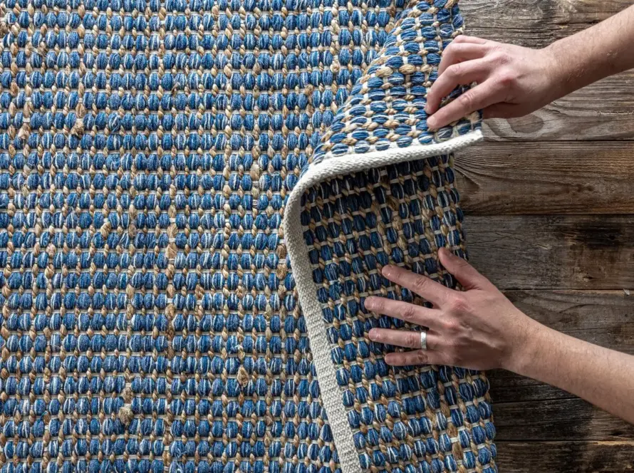 A person unrolling a woven rug with a blue and beige striped design, displaying its intricate texture on a wooden floor.