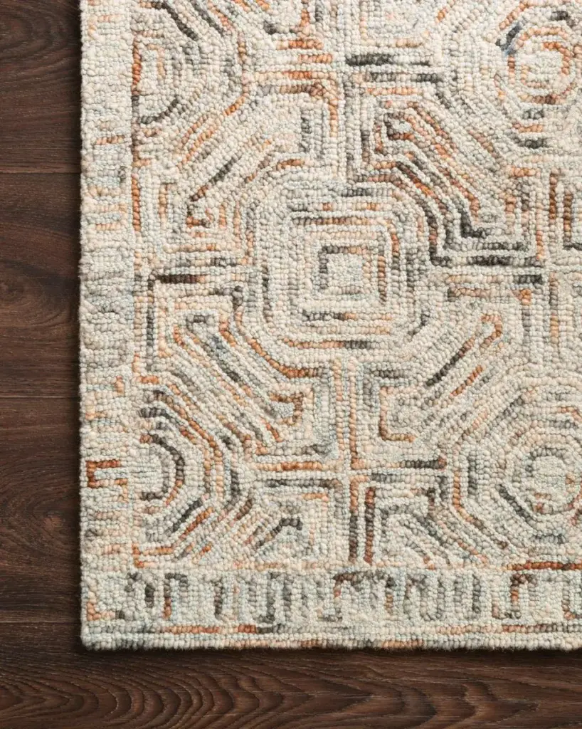 A close-up of a textured rug with an abstract geometric pattern in neutral tones, laid on a dark wooden floor.